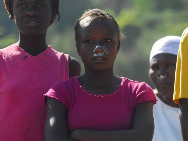 The image shows three young girls standing next to each other in front of a group of people, with a...