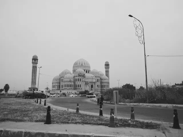 The image shows a black and white photo of a mosque in the middle of a street, surrounded by...