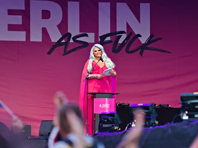The image shows a woman standing at a podium in front of a crowd. She is wearing a pink dress and...