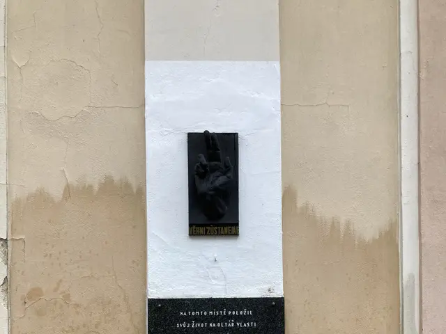The image shows a plaque on the side of a building with a hand on it. The plaque is black in color...