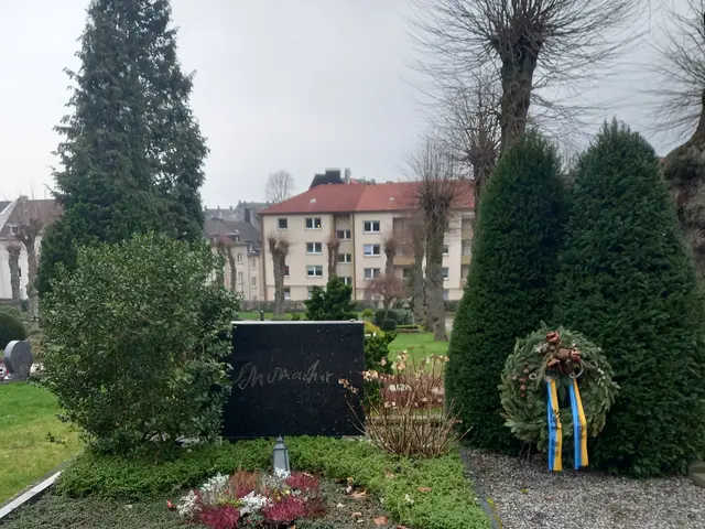 The image shows a memorial in the middle of a park with a wreath on it, surrounded by grass,...