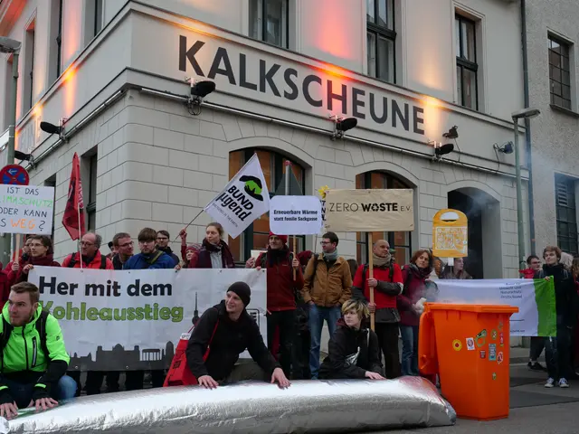 The image shows a group of people standing in front of a building, holding banners and placards...
