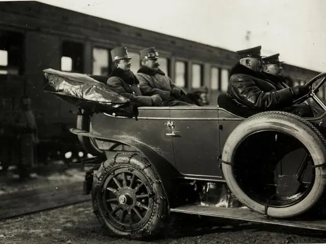 The image shows an old black and white photo of a group of men sitting in an old car, wearing caps,...