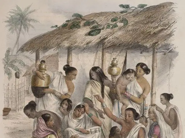 The image shows a group of women sitting around a table in front of a hut, with one woman holding a...