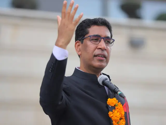 The image shows a man in a black suit and tie, wearing spectacles and a garland around his neck,...