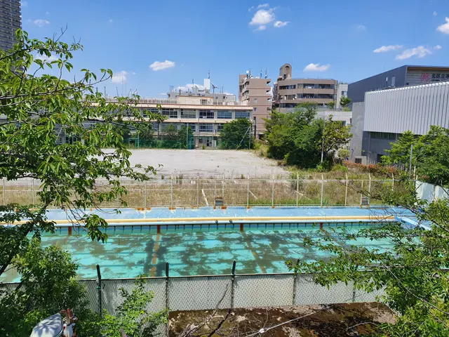 The image shows an abandoned swimming pool in the middle of a city, surrounded by a fence and...