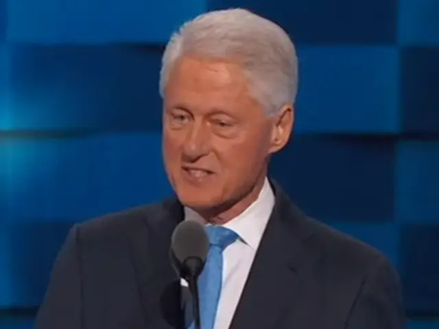 The image shows a man in a suit and tie standing at a podium with a microphone in front of him and...