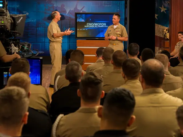 The image shows two men in military uniforms standing in front of a crowd of people sitting in...