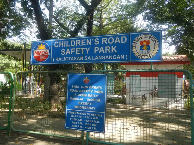 The image shows a children's road safety park with a metal fence surrounding it, two blue boards...