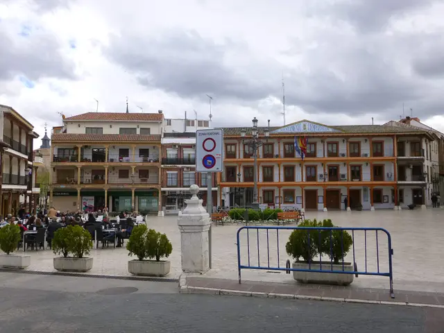 The image shows a bustling plaza mayor in the center of a city, with a group of people sitting on...