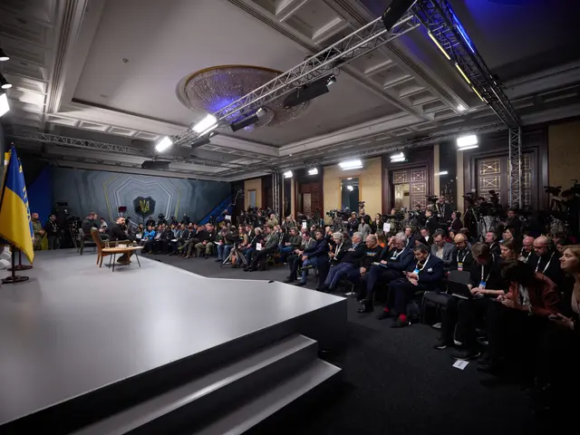 The image shows a large group of people sitting on chairs in front of a stage, with a table in the...