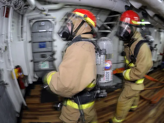 The image shows two firefighters in protective gear, wearing helmets and gas masks, walking through...