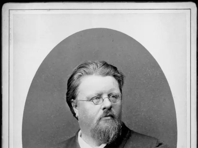 The image shows a black and white photo of a man with a beard wearing a coat and spectacles. At the...