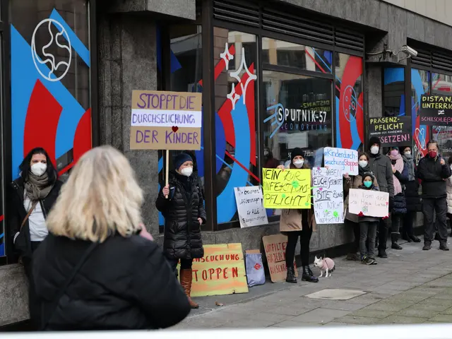 The image shows a group of people wearing masks and holding signs in front of a building with glass...