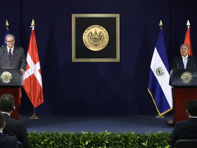 The image shows two men standing at podiums in front of a crowd of people sitting in chairs. The...