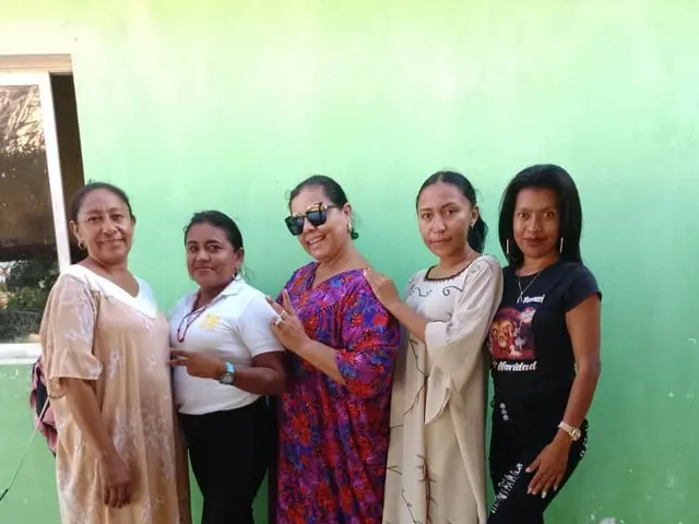 The image shows a group of women standing next to each other in front of a green wall, with a...