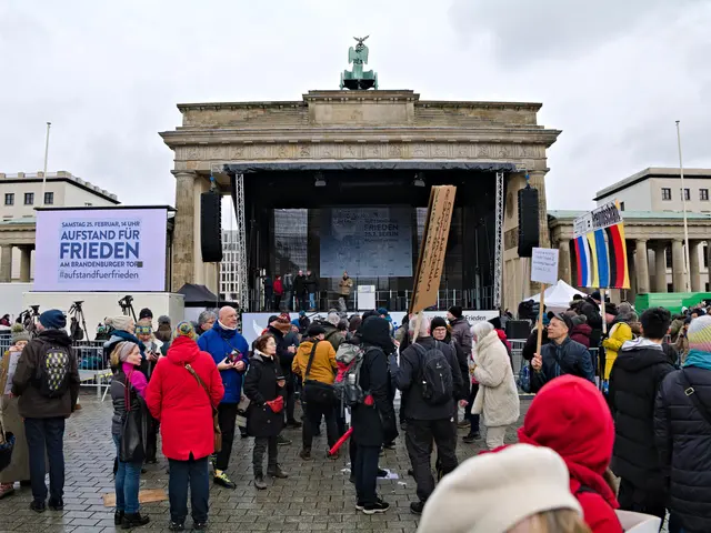 The image shows a crowd of people standing in front of a building with a stage in the center. On...