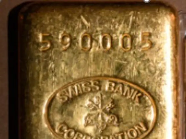 The image shows a gold bar in a plastic case on a white background. The bar is inscribed with text...