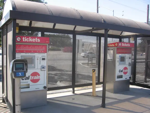 The image shows a bus stop with two ticket machines sitting on the side of it. In the background,...