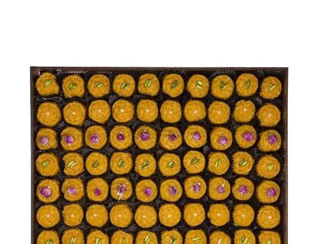 The image shows a box filled with a variety of yellow and pink sweets, such as kaju katli, on top...