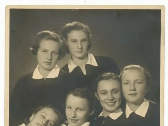 The image shows a black and white photo of a group of young girls in school uniforms, smiling and...