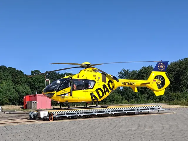 The image shows a yellow helicopter sitting on top of an airport tarmac, surrounded by trees and a...