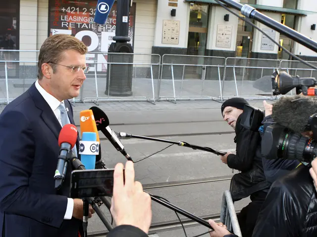 The image shows a man in a suit and tie standing in front of a group of reporters, some of whom are...