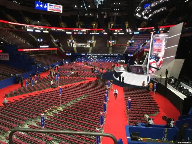 The image shows a large auditorium filled with lots of red chairs and a red carpet. There are a few...