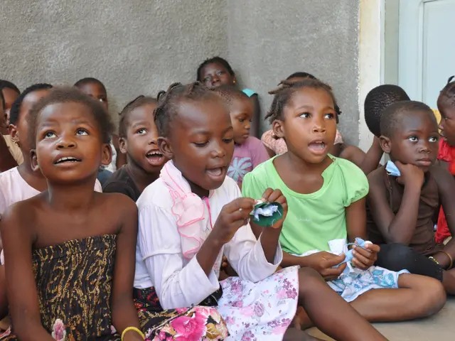 The image shows a group of children sitting on the floor in front of a building, with a wall and a...