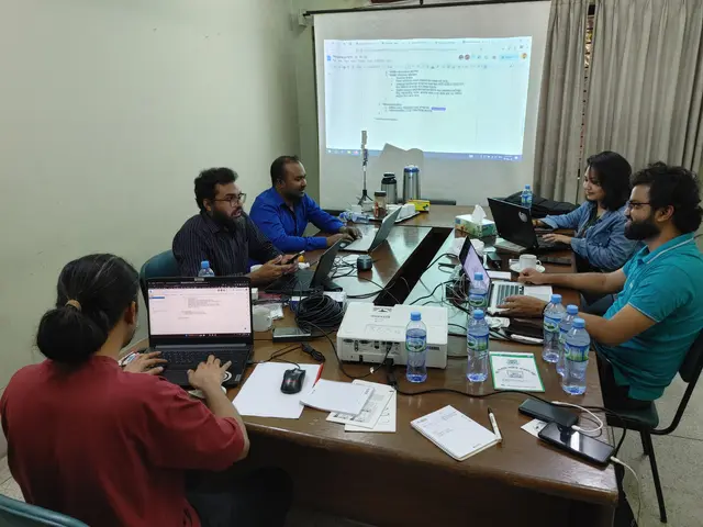 The image shows a group of people sitting around a table with laptops, bottles, papers, books,...