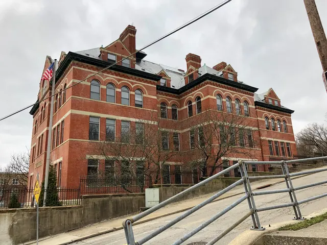The image shows a large brick building with a flag on top of it, surrounded by a metal fence, a...