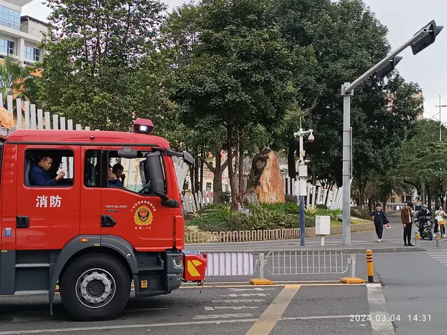 The image shows a red fire truck driving down a street next to a traffic light. There are people...