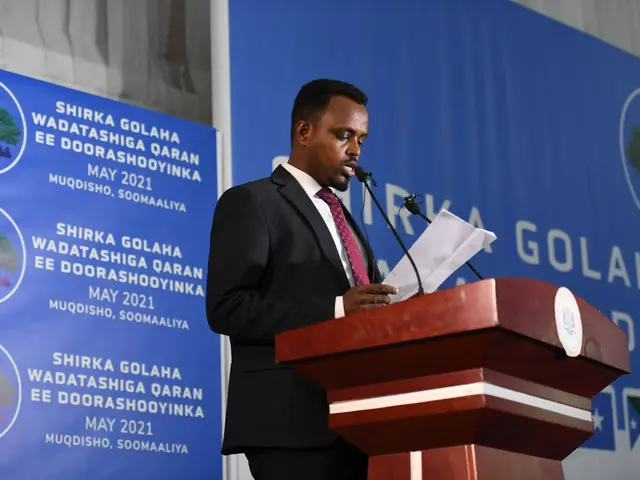 The image shows a man in a suit and tie standing at a podium, holding papers in his hand and...