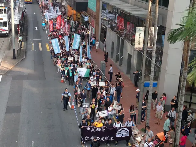 The image shows a large group of people walking down a street, holding banners and placards with...