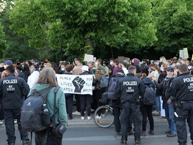 The image shows a large group of people standing on the side of a road, some of them holding...