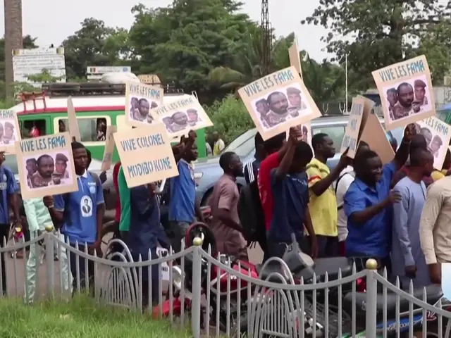 The image shows a group of people holding signs in front of a fence, surrounded by vehicles, trees,...