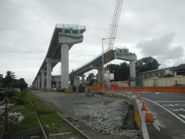 The image shows a construction site with a bridge in the background, a road with traffic cones on...