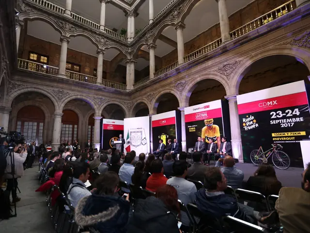 The image shows a group of people sitting in chairs in front of a crowd of people, some of whom are...