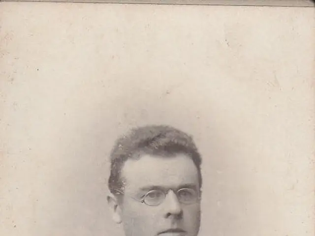 The image shows an old black and white photo of a man wearing a priest's robes and spectacles. At...