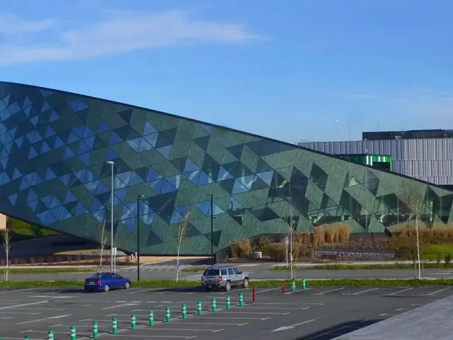 The image shows a large building with a lot of cars parked in front of it, surrounded by street...