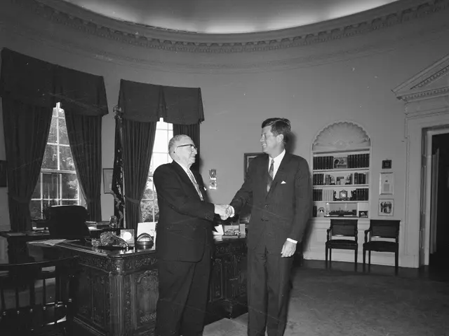 The image shows two men shaking hands in an oval office. They are both wearing blazers, ties, and...