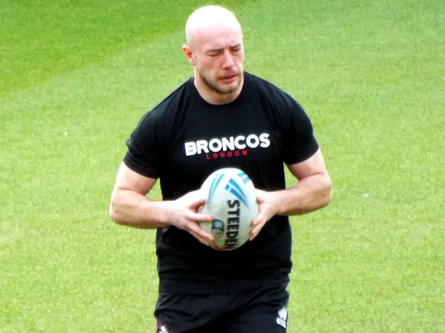 The image shows a man running with a rugby ball in his hands on a grassy field. He is wearing a...