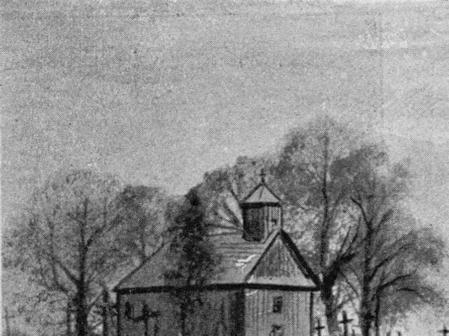 The image shows a black and white photo of a small church in the middle of a field, surrounded by a...