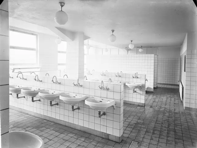 The image shows a black and white photo of a public restroom with sinks, taps, walls, lights, and...