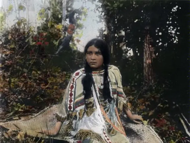 The image shows an old photo of a Native American Indian girl sitting on a rock in the woods,...
