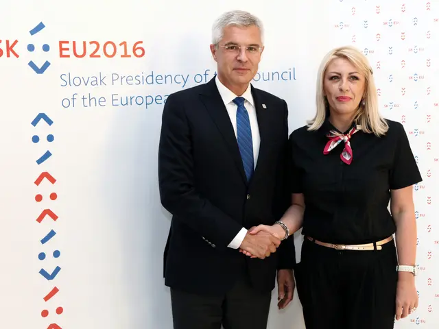 The image shows a man and woman standing next to each other in front of a wall, both of them...