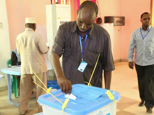 The image shows a man putting a voting paper in a blue and white box, surrounded by a group of...