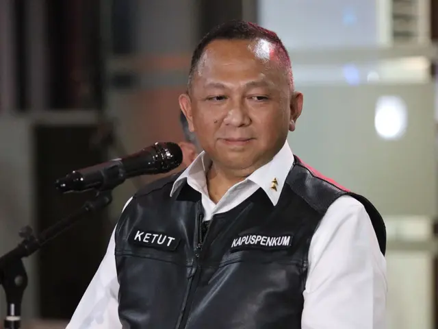 The image shows a man in a black vest and white shirt standing in front of a microphone, with a...