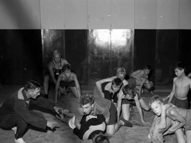 The image shows a group of young boys wrestling in a gym, with a wall in the background. The image...