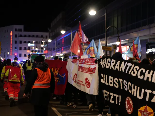The image shows a group of people walking down a street at night, holding flags and banners with...